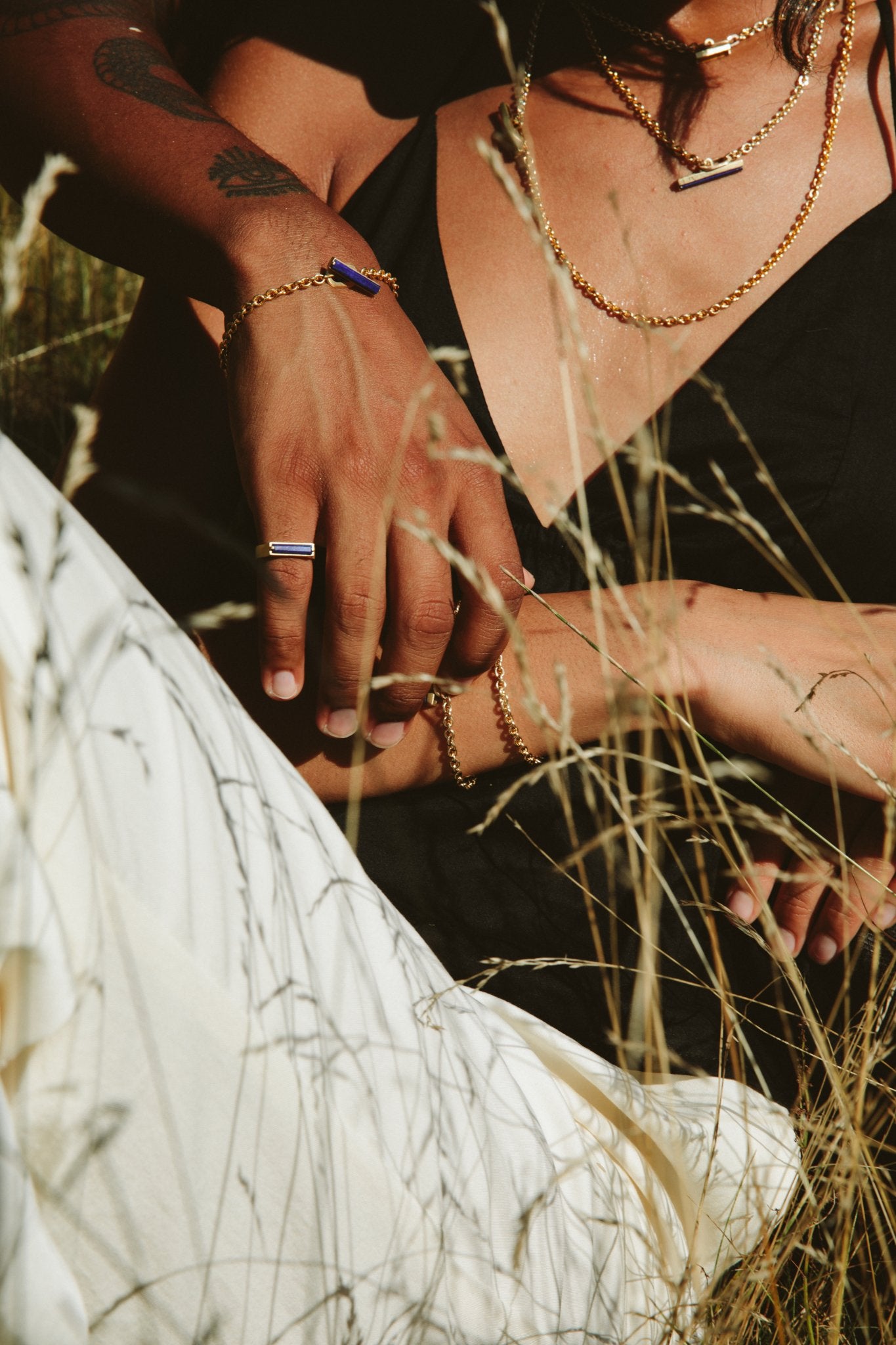 A couple embracing one another in wild grass, wearing MEILI Pillar Gemstone Rings and MEILI Pillar Toggle chain in yellow gold. 