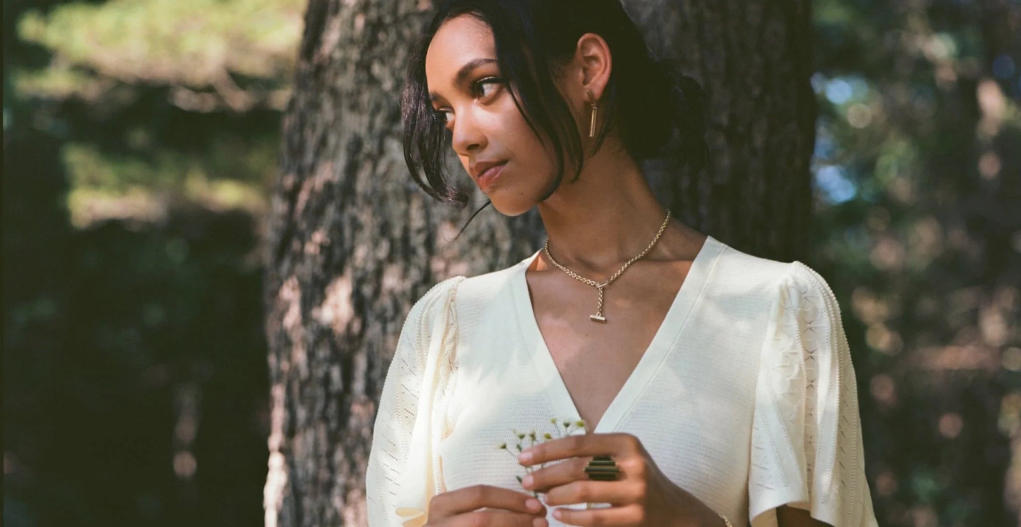A women holding wildflowers stands in nature wearing a gold toggle clasp necklace, and Pillar 5 stone ring from Meili Pillars fine jewelry collection